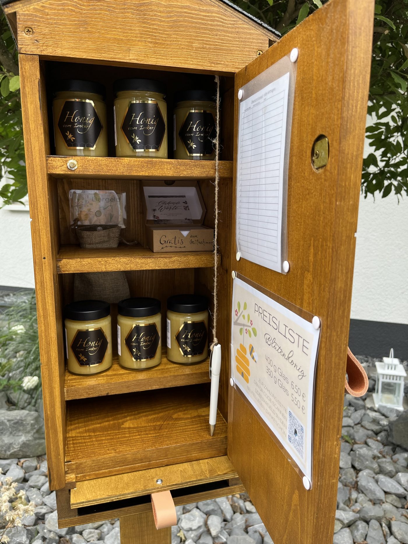 Geöffneter Ehrenkasten aus Holz mit Honiggläsern „Honig vom Imker“, Preisliste mit QR-Code und Stift an der Tür