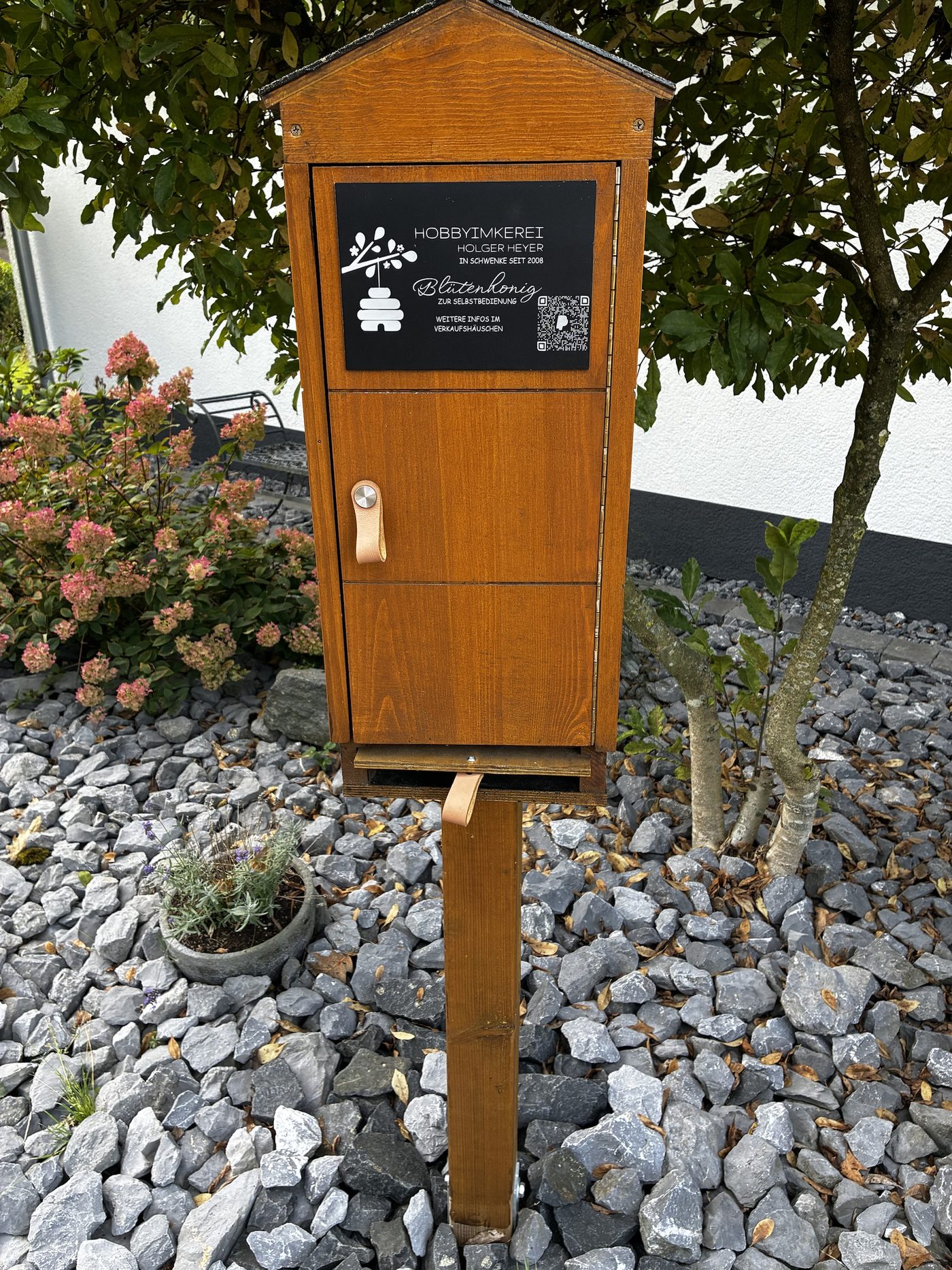 Der Ehrenkasten von vorn: Holzhäuschen auf einem Pfosten mit schwarzem Schild Hobbyimkerei Holger Heyer, Blütenhonig zur Selbstbedienung