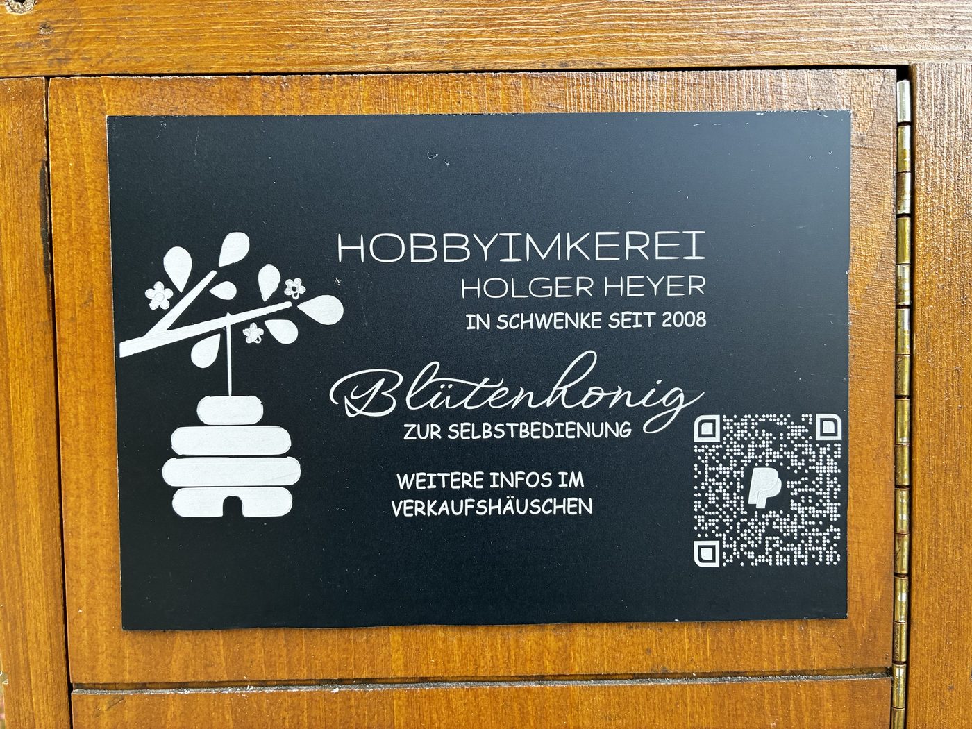 Schwarzes Schild am Ehrenkasten: Hobbyimkerei Holger Heyer in Schwenke seit 2008, Blütenhonig zur Selbstbedienung, mit PayPal-QR-Code